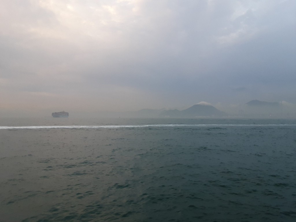 View of the Inter-island Sea near Peng Chau, Hong Kong on a hazy day. The trail of an unseen boat draws a horizontal line across the frame. A cargo boat floats in the distance. Further away is a foggy shore; a mountain peaks through the clouds. Reference image for Yip Kai Chun's 2025 solo exhibition 'How to draw a line across the sea?'.