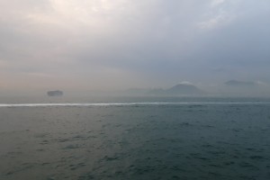View of the Inter-island Sea near Peng Chau, Hong Kong on a hazy day. The trail of an unseen boat draws a horizontal line across the frame. A cargo boat floats in the distance. Further away is a foggy shore; a mountain peaks through the clouds. Reference image for Yip Kai Chun's 2025 solo exhibition 'How to draw a line across the sea?'.